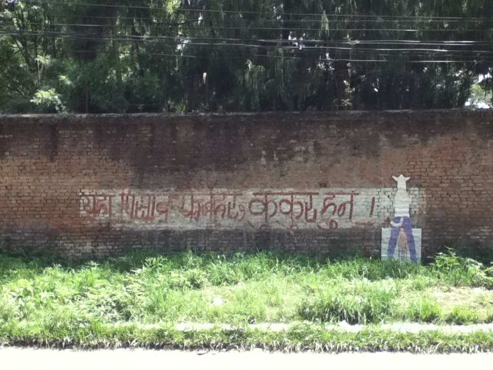 a wall with writing on it that says, in Nepali, 'Those who urinate here are dogs.' and a poster of a half-human-half-dog creature urinating